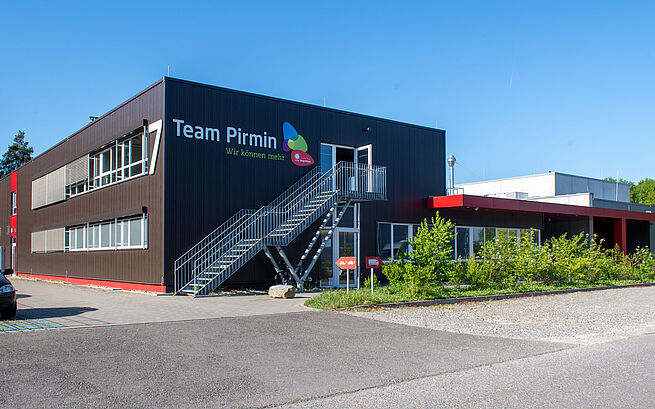 Modernes Firmengebäude mit der Aufschrift „Team Pirmin – Wir können mehr“ bei Sonnenschein.