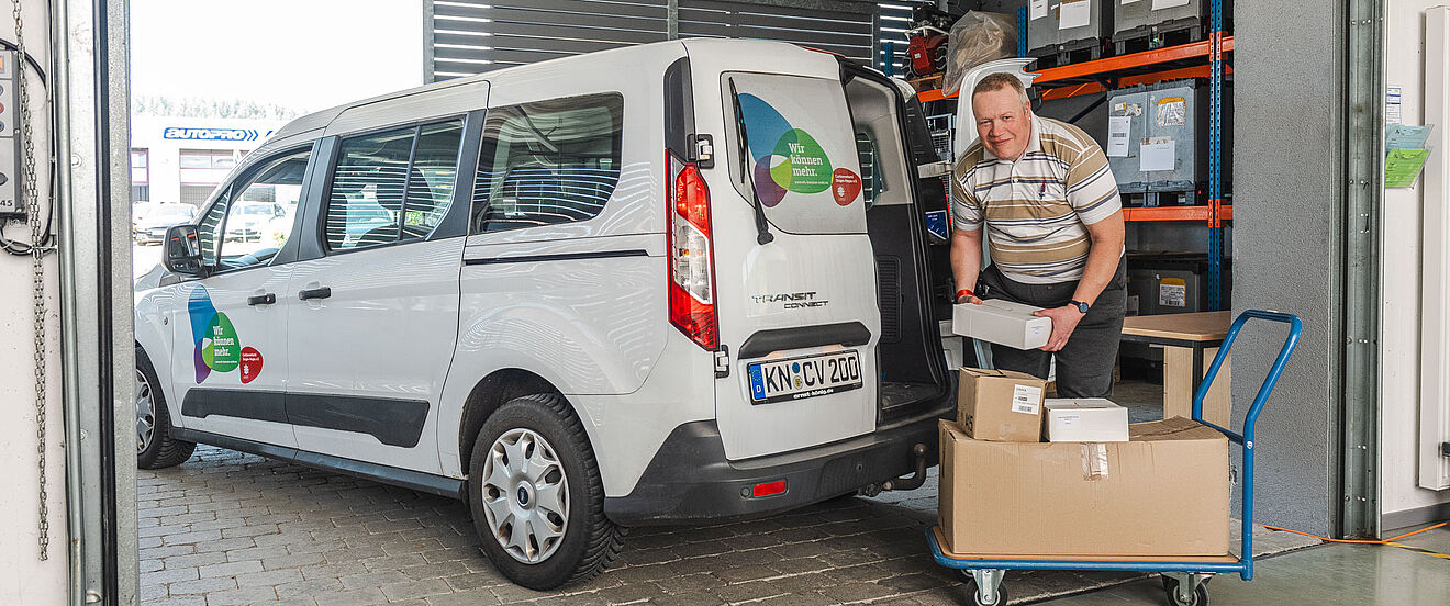 Ein älterer Mann lädt Pakete aus einem weißen Lieferwagen vor einem Lagerhaus.