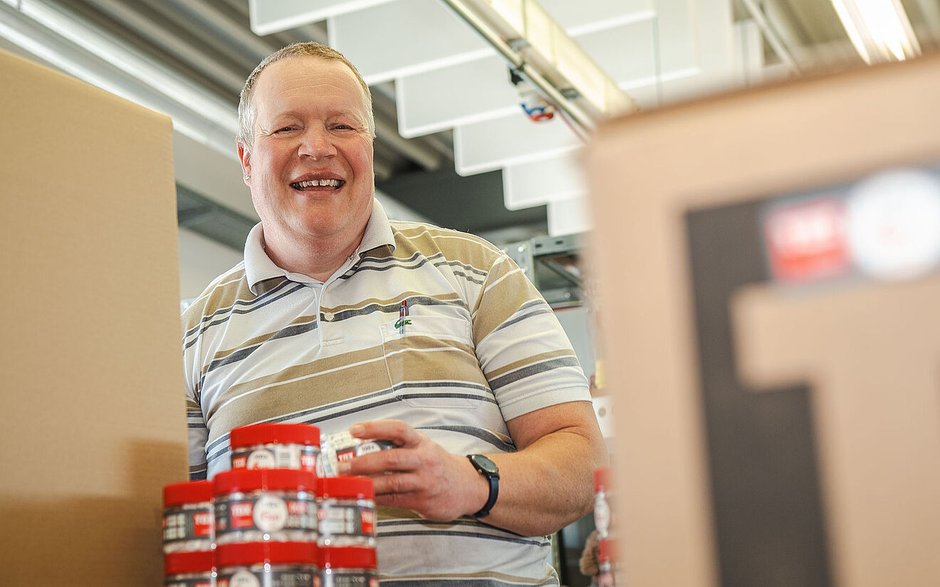Lächelnder Mann verpackt rote Dosen in Kartons in einer Werkstattumgebung.