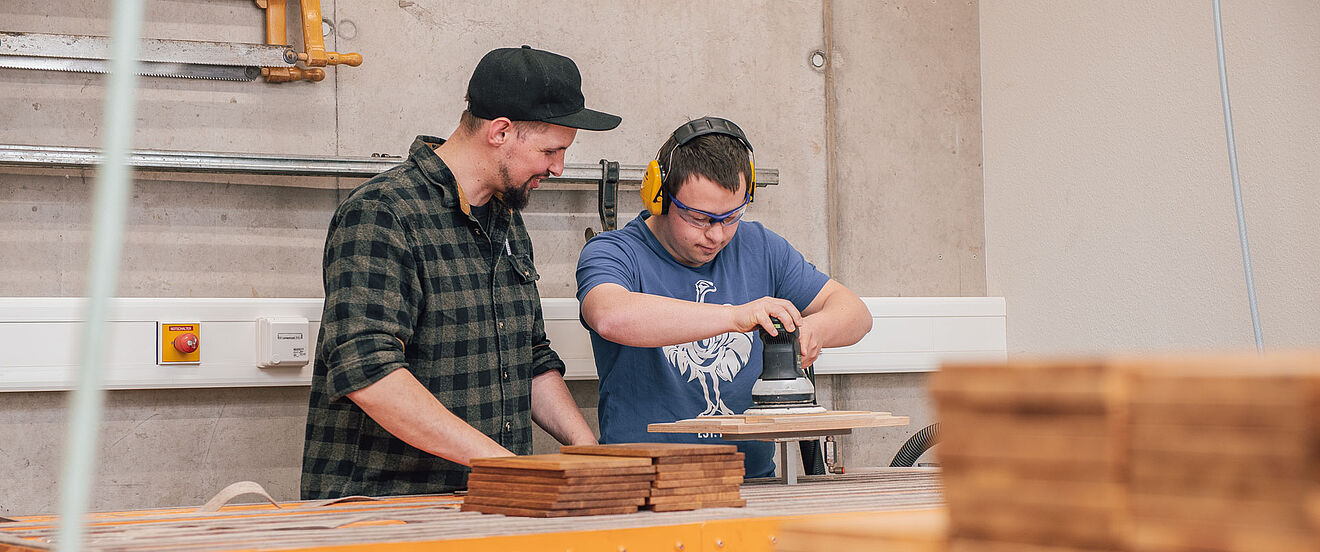 Zwei Männer schleifen gemeinsam ein Holzbrett in einer Werkstatt.