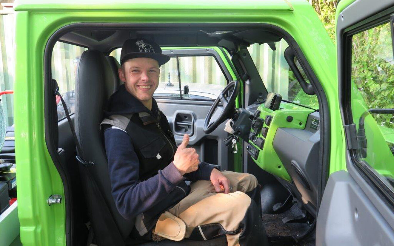Ein lächelnder Mann im Arbeitsoutfit sitzt in einem grünen Fahrzeug und zeigt den Daumen hoch.