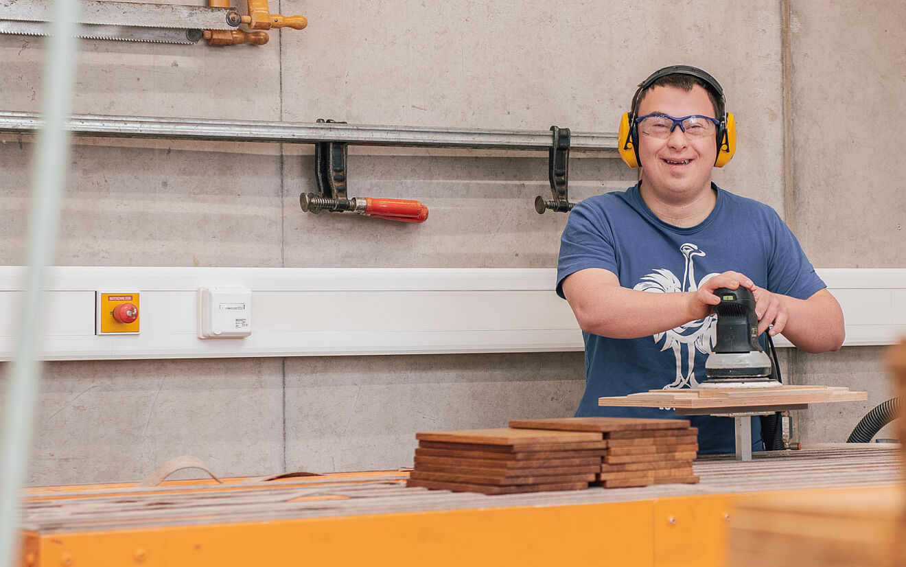 Mann mit Gehörschutz schleift Holzplatten in einer Werkstatt mit großer Schleifmaschine.