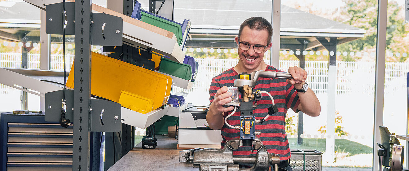 Ein Mann mit Brille arbeitet lächelnd an einer Werkbank mit einer Maschine.