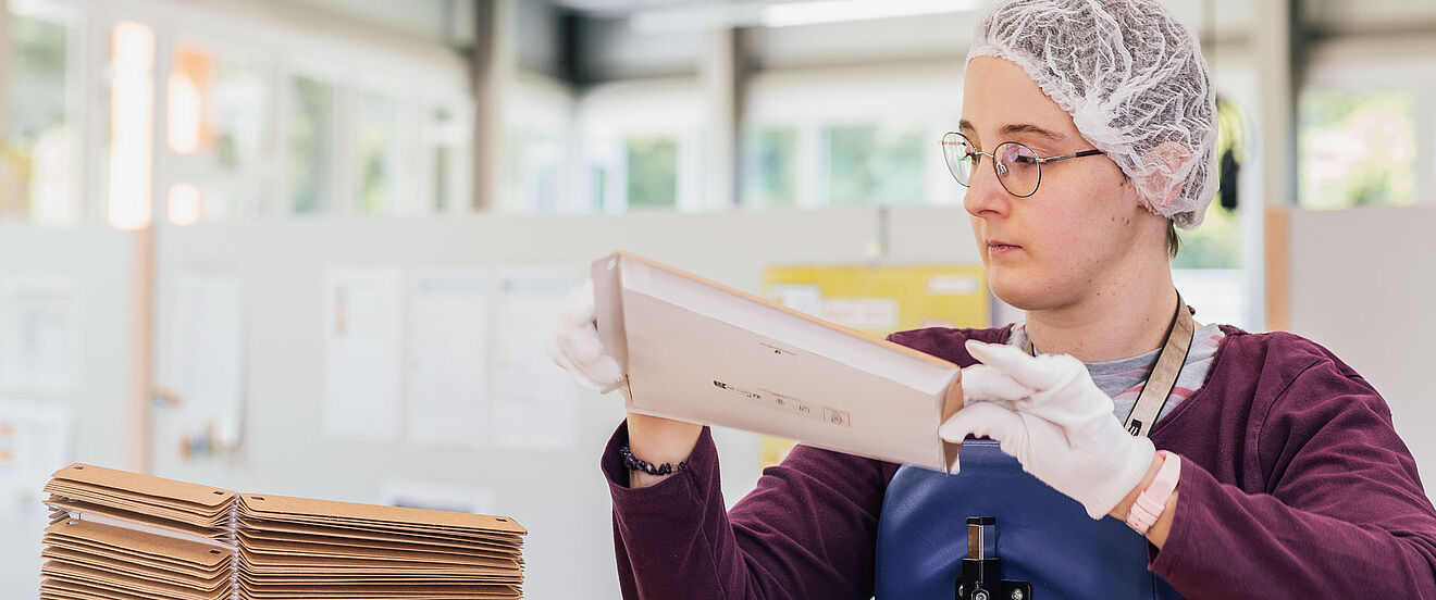 Eine Frau mit Haarnetz und Handschuhen faltet Verpackungen an einem Arbeitstisch.